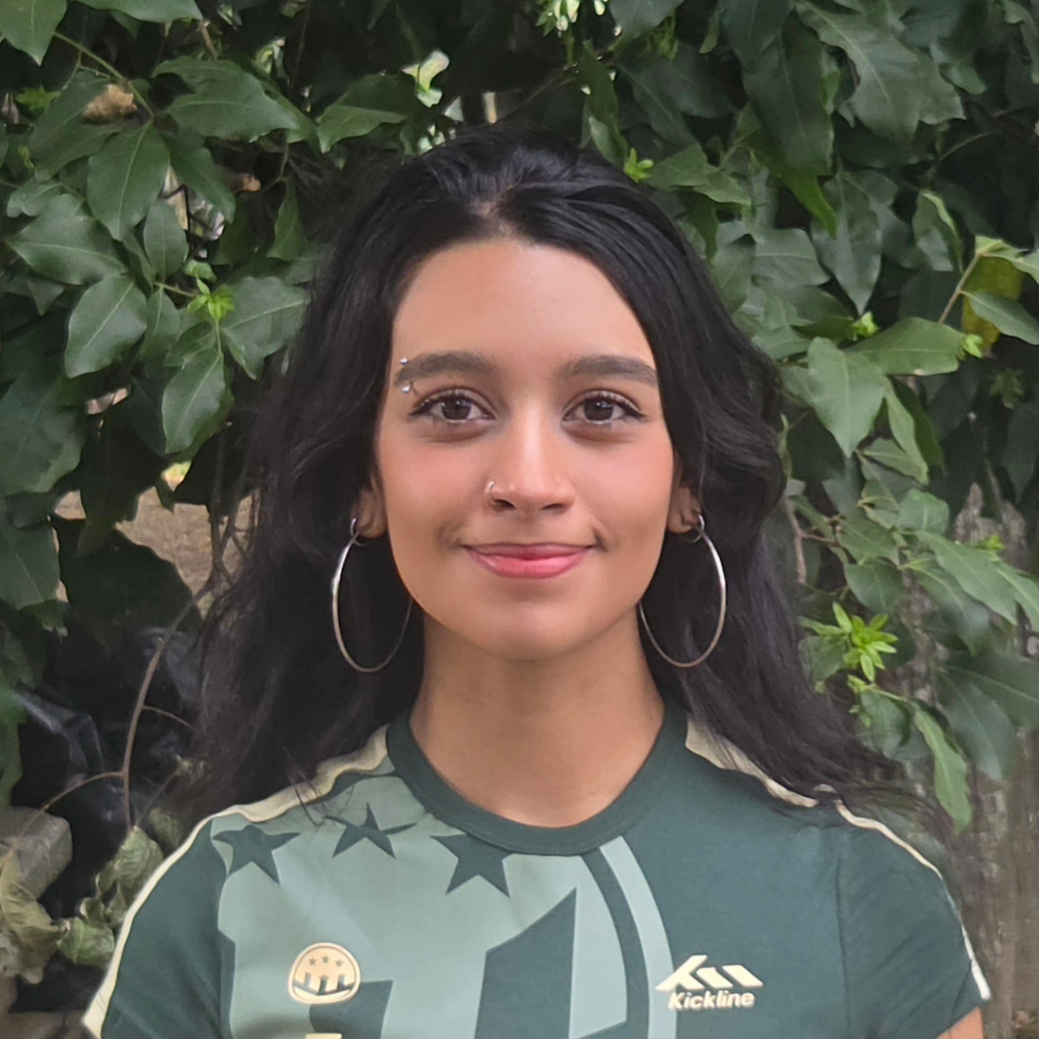 A brown woman wearing a blue-green football (soccer, for Americans) jersey straight ahead at the camera and smiles. Her hair is long, black, and lightly wavy. She has two sets of hoops on her ears, one small, the other very large; a ring in her right nostril; and a curved bar piercing in her right eyebrow. Her eyes are brown. She stands in front of a plant, possibly a thick patch of ivy or a shrub.
