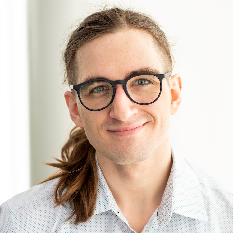 A white person standing and smiling against a bright, mostly white background. They are wearing glasses and a white dress shirt with small dots. Half of their face is lit.