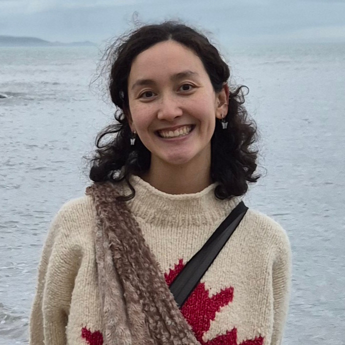 A woman of white and East Asian descent smiles wide into the camera. She has curly brown hair, brown eyes, and freckles. She is wearing earrings, a white knitted sweater with a red Canadian maple leaf, and two cross-body bags. The background is a hazy ocean scene, with small, choppy waves and hills in the far distance.