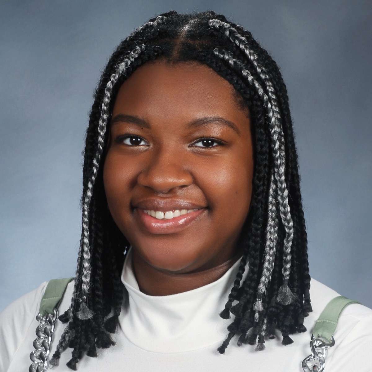 A Black woman with a round face is smiling at the camera in front of a blue-gray background. She wears her hair in braids, some of which are silver. She is wearing a white shirt under a sea green and silver layer.