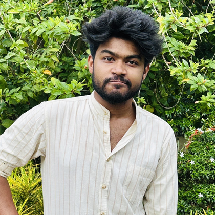 A South Asian man with thick black and gray hair standing up looks dead at the camera with tight lips. He is wearing a patially buttoned striped cream shirt, and his hand is on his hip (hip not visible). In the background are mostly green plants, with some white and red flowers.