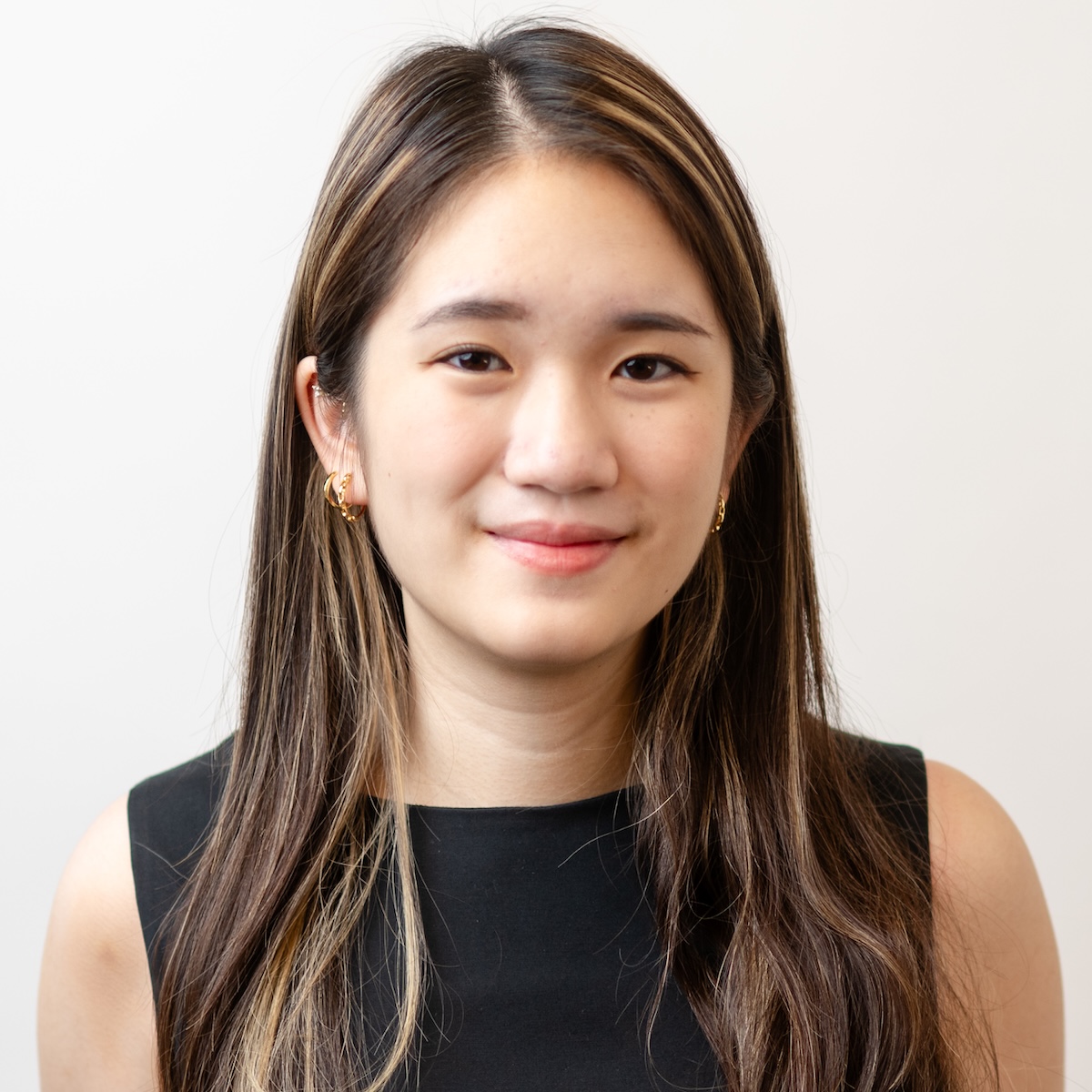 A Southeast Asian woman with brown eyes and long brown hair streaked with blonde gives a slight smile to the camera. Standing in front of a white background, she is wearing several gold earrings and a black sleeveless top.