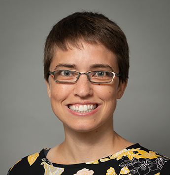 A white woman is giving a wide smile. She stands against a plain, gray background. She is wearing glasses and a black and yellow top.