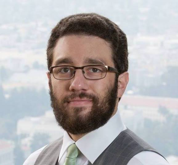 A white man with a beard and short, curly hair. He is wearing a white shirt, suit vest, green tie, and glasses, and he is slightly smiling at the camera. The background faintly shows some hills and trees.