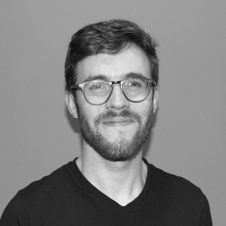 A grayscale image of a white man wearing a t-shirt and glasses. His eyes are creased as he gives a tight-lipped smile. His hair is short on the sides and a bit longer on top; he is sporting a short beard.