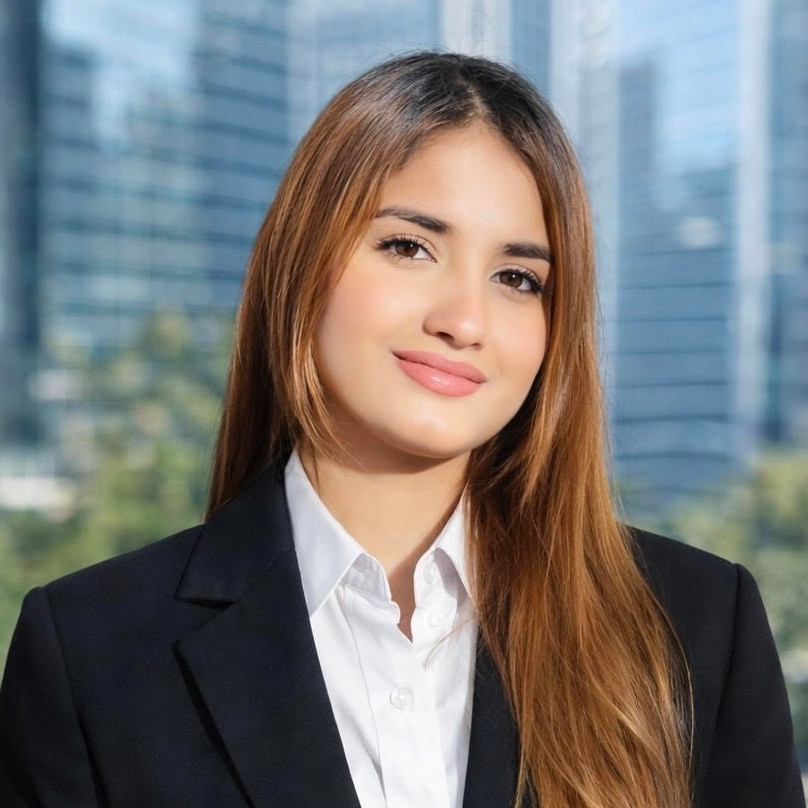 A South Asian woman with brown eyes and brown hair (highlighted a more auburn hue) looks at the camera with a tilted head and a small, confident smile. She is wearing a white button-up shirt with the top button unbuttoned and a black blazer. In the background are high-rise buildings and, below them, trees.