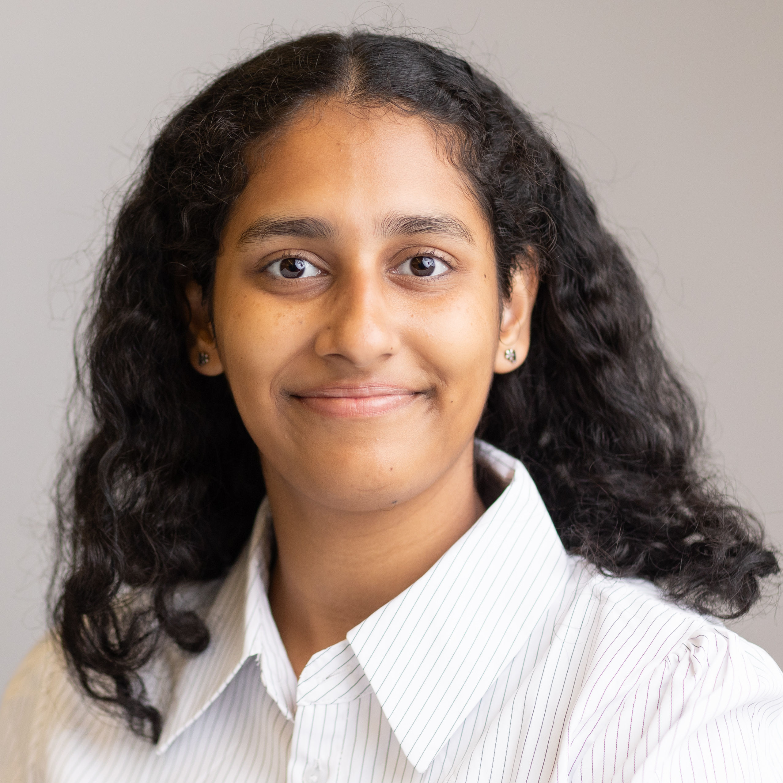 A South Asian woman with curly, wavy black hair gives a slight smile to the camera. She is wearing a striped white dress shirt against a light gray background.