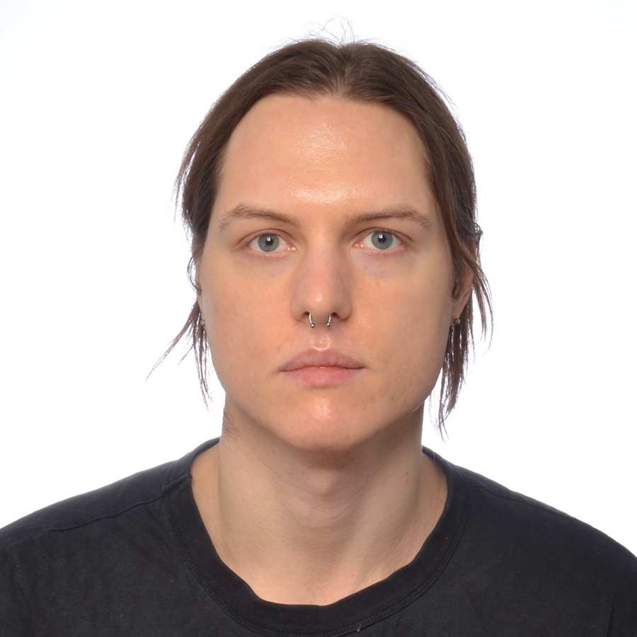 A white person with steel-blue eyes looks straight at the camera with a neutral expression. His hair is straight and parted in the middle, with wisps going in different directions on both sides of his head. He has a septum piercing with a circular barbell ring and is wearing a black t-shirt. The background is uniform white.